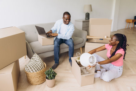 Happy african american couple unpacking belongings from cardboard boxes in new apartment, starting...