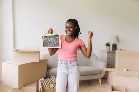 Excited african american woman showing moving day sign while celebrating moving into new apartment surrounded by cardboard boxesの写真素材