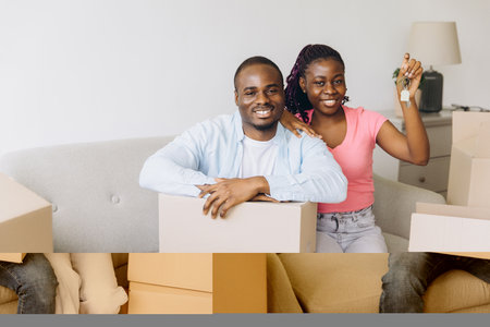 Excited couple showing keys to their new apartment while sitting on sofa surrounded by cardboard boxes, happy to start new life chapterの写真素材