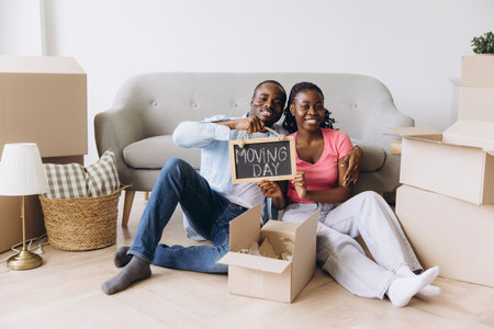 Smiling african american couple sitting on floor among cardboard boxes showing moving day sign in new apartmentの写真素材