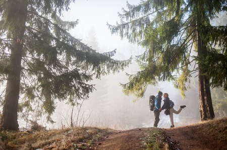 Beautiful couple with backpacks is having fun on foggy autumn day in the mountain forest. Side view of man and woman holding hands.の写真素材