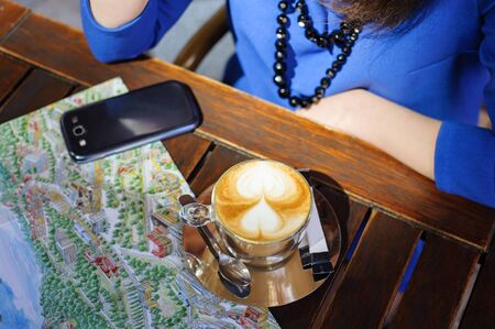 Cropped image young girl sitting at the table with cup of coffee, and smart phone and reads the mapの写真素材