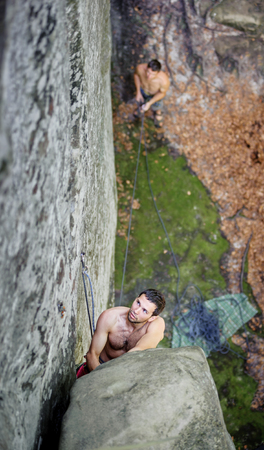 Young rock-climber climbing over big boulder on steep cliff, his partner belayingの写真素材