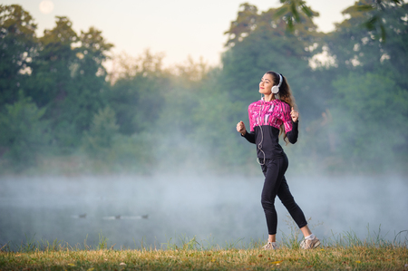 Fullbody portrait of beautiful woman runner on foggy morning standing near lake with headphones on.の写真素材
