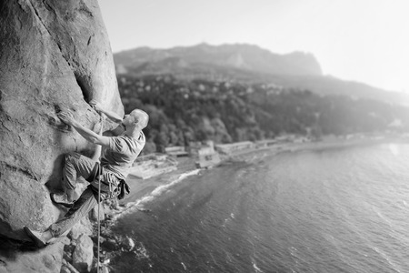 Side view of young man rock climber making difficult move up on multi-pitch against scenic sea coast background. Climbing equipment. Healthy lifestyle concept. Panoramic picture. Black and whiteの写真素材
