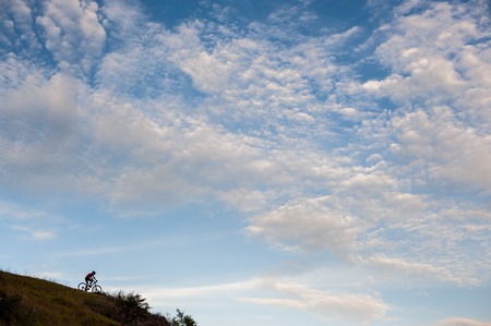Silhouette of a cross country cyclist going downhill against cloudy blue sky.の写真素材