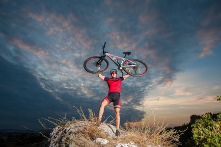 Young happy mountain biker celebrating making the top of the peak. Cyclist is holding his bike up.の写真素材