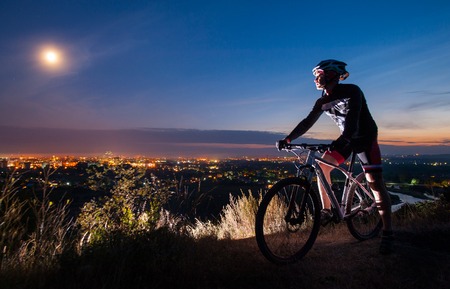 Young athlete biker with mountain bike on top of the hill observing the evening city view.の写真素材