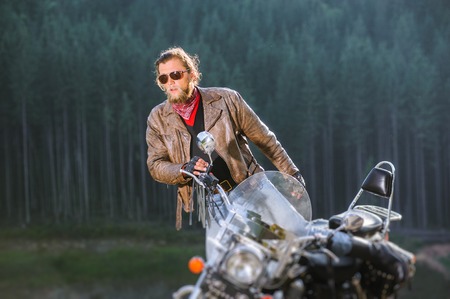 Handsome young serious biker is sitting down on his long distance travel motorbike with forest on...