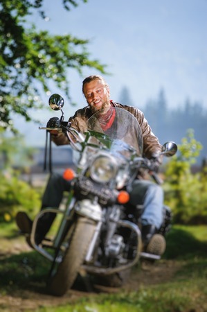 young biker with beard driving his cruiser motorcycle in the forest and smilling. Man is wearing leather jacket and blue jeans.  Tilt shift lens blur effectの写真素材