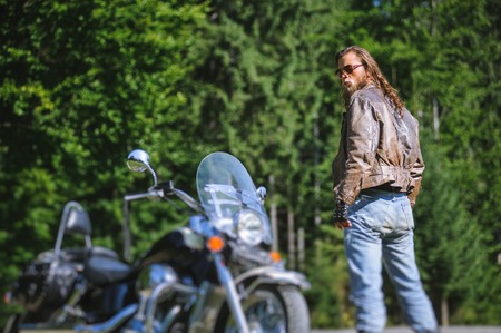 Handsome brutal man with long hair and beard standing near his custom made cruiser motorcycle. Biker is wearing leather jacket and sunglasses on sunny day. back view. tilt shift soft effectの写真素材