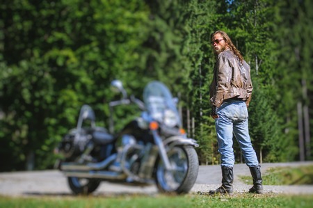 Handsome man with long hair and beard standing near his custom made cruiser motorcycle. Biker is wearing leather jacket and sunglasses on sunny day. tilt shift soft effectの写真素材