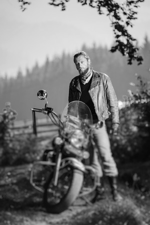 Young handsome biker standing by his custom made cruiser motorcycle on a sunny day with forest on the background. Tilt shift lens blur effect. Black and whiteの写真素材