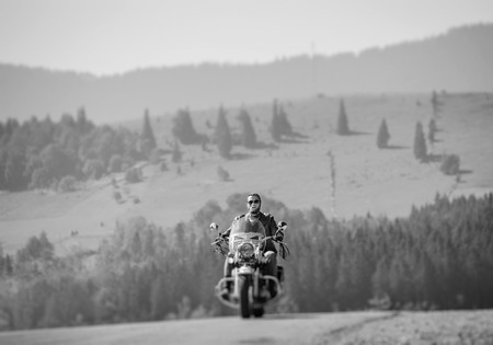 Handsome biker in sunglasses on mountainous road, man is riding his custom made travel motorbike. Sunny day in the mountains. Tilt shift soft effect. Black and whiteの写真素材