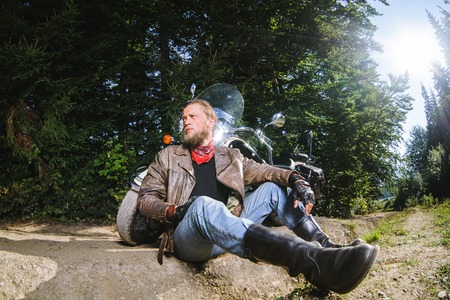 Bearded biker posing with his shiny stylish custom made cruiser motorbike. Biker is sitting on the ground near his bike wearing leather jacket, gloves and boots. Looking to the distanceの写真素材