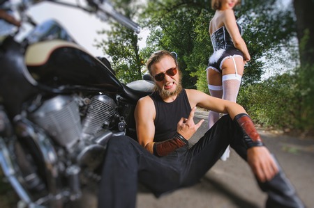 Sexy couple of bikers. Bearded biker sitting on the ground near his bike and giving the devil horns gesture. Attractive  woman wearing costume of bunny standing behind the man. Tilt lens blur effectの写真素材