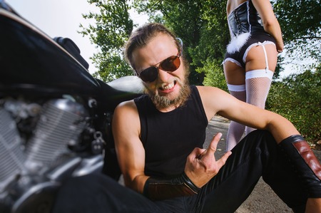 Close-up portrait of hardcore bearded biker sitting on the ground near his bike and giving the devil horns gesture. Attractive girl wearing costume of bunny. Tilt shift lens blur effectの写真素材