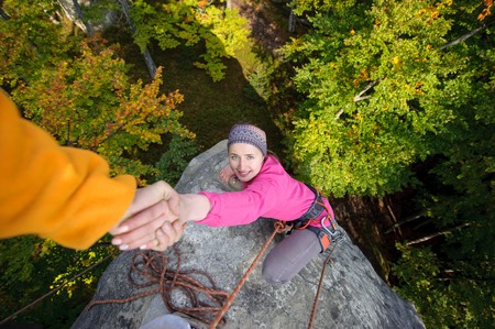 Rockclimber is helping a young beautiful girl to climb to the peak of mountain. Man giving a hand to the woman. Warm autumn eveningの写真素材