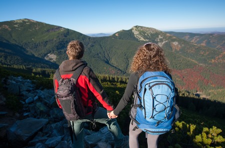 Couple of hikers standing at the mountain peak holding hands on a warm sunny autumn day in the mountains. Young couple looking into distance enjoying scenic nature landscape. Rear viewの写真素材