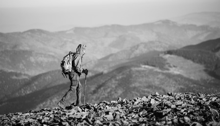 Male sportsman backpaker walking on the rocky mountain ridge with beautiful mountains on background. Man is wearing jacket and has trekking sticks and backpack on. Sunny day. black and whiteの写真素材