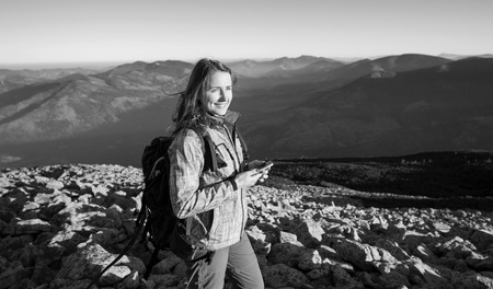Portrait of young cute woman backpacker standing on rocky top of the mountain, looking at the sun and holding cell phone. Ecotourism and healthy lifestyle concept. Sunny evening. black and whiteの写真素材