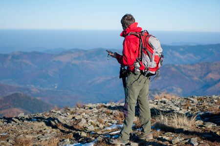 Portrait of young man backpacker standing on the peak of the mountain and looking at gps coordinates. Beautiful mountains on the background. Rear view, sunny autumn day.の写真素材