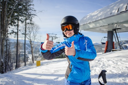 Portrait of young happy woman skier at ski resort smiling and showing thumbs up. Winter sports concept. Woman is wearing blue jacket and blue pants, helmet and orange goggles.の写真素材