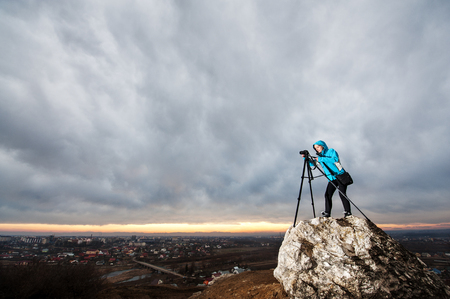 Female photographer is standing with her camera on tripod on the big rock at city overview point. Woman is taking picture of the cityscape. Dark blue evening sky in the backgroundの写真素材