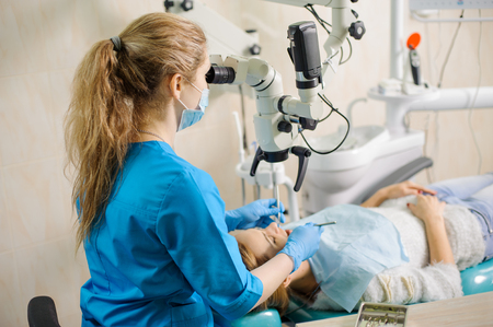 Young female dentist treating root canals using microscope at the dental clinic. Young woman patient lying on dentist chair with open mouth. Dentist wearing mask and glovesの写真素材