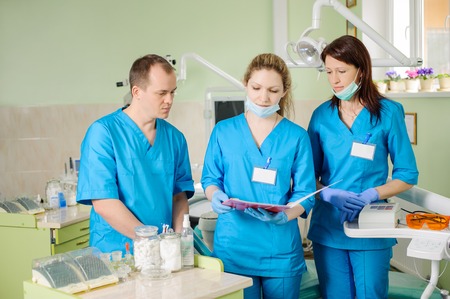 Three dentists looking at the patient folder at the stomatology clinic. Doctors wearing masks and gloves. Dental care for people. Stomatology equipmentの写真素材