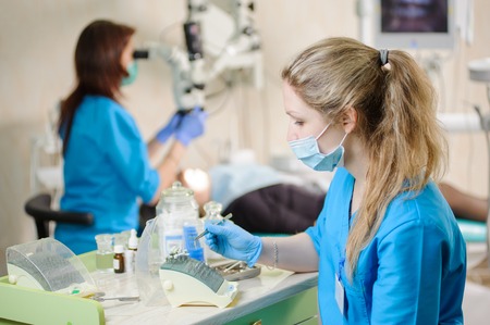 Young female dentist assistant taking dental drill on foreground. Doctor treating caries using...