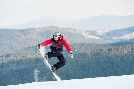 Man boarder jumping on his snowboard and taking his for the edge on top of a mountain against the backdrop of mountains, hills and forests in the distance. Bukovel, Carpathian mountainsの写真素材
