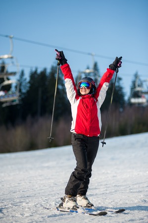 Portrait of female skier wearing helmet, red jacket and ski goggles standing on snowy slope with hands raised up in sunny day with forest and blue sky in background.の写真素材