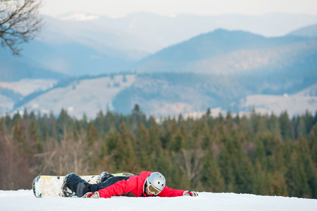 Carefree snowboarder wearing helmet, red jacket, gloves and pants lying on ski slope on the background of beautiful mountains and forests, winter sports concept. Carpathian mountains, Ukraineの写真素材