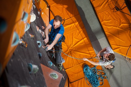 Strong man practicing climbing on rock wall indoors, view from above. Man standing on the ground insuring the climberの写真素材