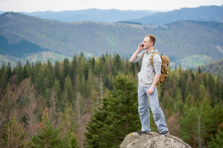 Male hiker with backpack standing on top of a mountain looking away and talking on the phone on the blurred background of forest valley and hills.の写真素材