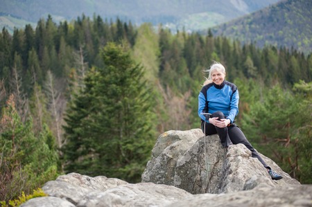 Smiling woman in a blue jacket with climbing equipment sitting on the top of the rock on the blurred background of forest valley.の写真素材