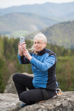 Tourist female in a blue jacket sitting on the rock makes the photo on the her phone with forest and mountains in blurred background. Close-upの写真素材