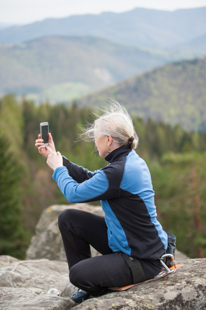 Hiker female in a blue jacket with climbing equipment sitting with back to camera on the rock makes selfie on the her phone with forest and mountains in blurred background. Close-upの写真素材