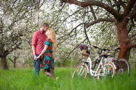 Romantic couple stand near the bicycles and tree holding hands in spring garden. Woman with long blond hair wearing flowered dress and man in a red shirt and jeansの写真素材