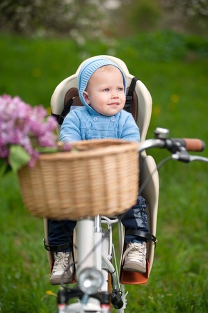 Little baby boy in blue jacket with a hood sitting in bicycle chair and looking away against the background of fresh greenery in spring gardenの写真素材