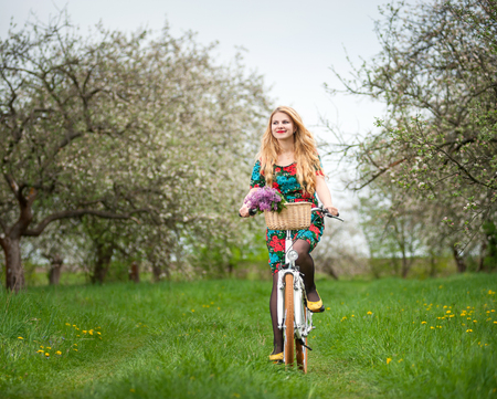 Beautiful woman with long blond hair wearing flowered dress and yellow shoes riding a white city bicycle with flowers basket in spring gardenの写真素材