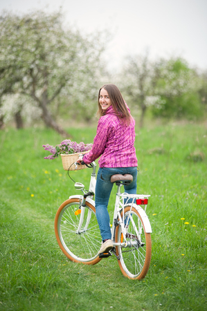 Brunette female girl wearing purple jacket and jeans and riding a vintage white bicycle with lilac flowers basket in spring garden. Rear viewの写真素材