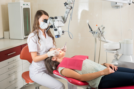 Female dentist working with microscope at modern dentist clinic. Doctor is wearing masks and gloves and treating cariesの写真素材