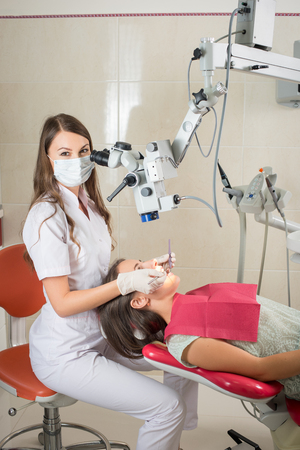 Woman dentist working with microscope and treating caries at modern dentist clinic. Doctor is wearing mask, gloves and looking at the cameraの写真素材