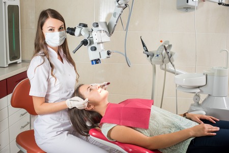 Female dentist working with microscope and treating tooth at modern dentist clinic. Doctor is wearing mask, gloves and looking at the cameraの写真素材