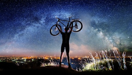 Silhouette of biker hold his bicycle above in the hands on the top of the mountain nearly city. Milky way and night city shining on the backgroundの写真素材