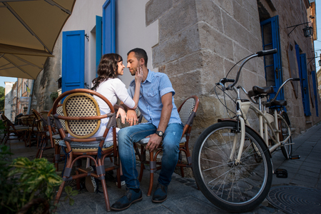 Loving couple sitting in sidewalk cafe near their tandem bike. Wife's hand gently concerns cheeks her husband, they tenderly looking at each otherの写真素材