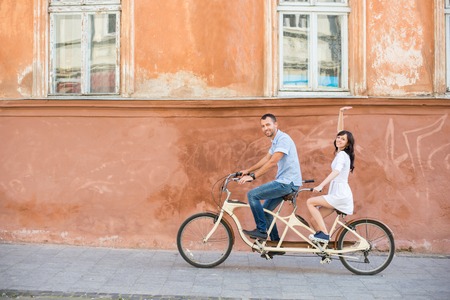 Happy couple riding on retro tandem bicycle at the street city against the background of the old orange wall with windows. The man runs a bicycle, a girl in white dress raised her hand up. Lvivの写真素材