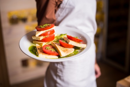 Close-up of woman in a white apron holding in hand delicious vegetarian lunches tomato salad Caprese with basil sauce and cheese on a white plate.の写真素材